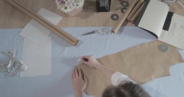 Masada kıvrımlı ve desenli çalışan moda tasarımcısının yavaş çekim videosu, bir sabun dilimi çizer, etrafında makas, santimetre, çizimli bir defter, dikiş stüdyosu, üst kattan görünüm