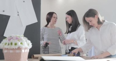 Üç moda tasarımcısının bir masada oturup tartıştığı yavaş çekim videoları, kıvrımlar ve desenler, makas kağıdı, akıllı telefondan bakan kızlar, dikiş stüdyosu, güneş ışığı, gülücükler.