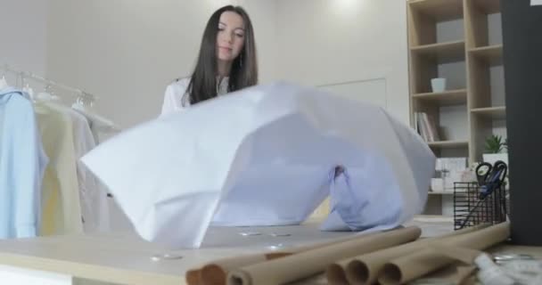 De jonge langharige brunette meisje spreidt stof op een tafel voor het maken van jurken in naaisalon, blauwe kleur, ambachtelijk papier — Stockvideo
