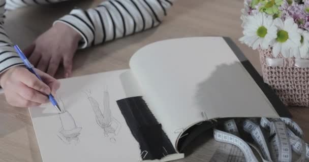 Langsom film av jenta tegner en skisse av den fremtidige modellen av klær i notisboken, verktøy for å sy, sy agenturet, en haug med blomster, papirhåndverk, oversikten – stockvideo