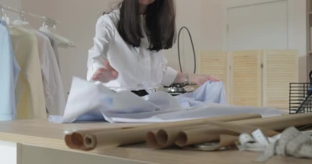 De jonge langharige brunette meisje spreidt stof op een tafel voor het maken van jurken in naaisalon, blauwe kleur, ambachtelijk papier — Stockvideo