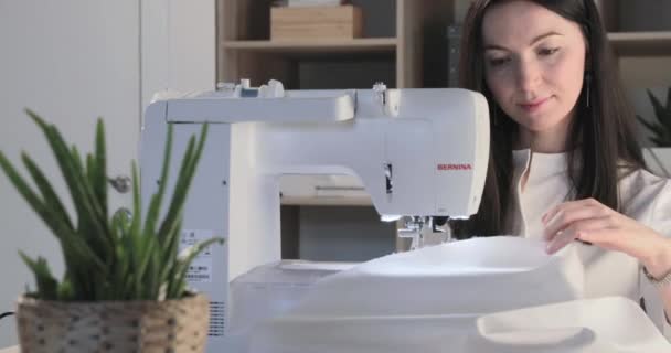 Russia, Saint-Petersburg, 01 March 2019: The beautiful young girl sits at the automatic sewing machine in sewing studio and sews some clothes, the brunette, long hair, flowers and accessories — Stock Video