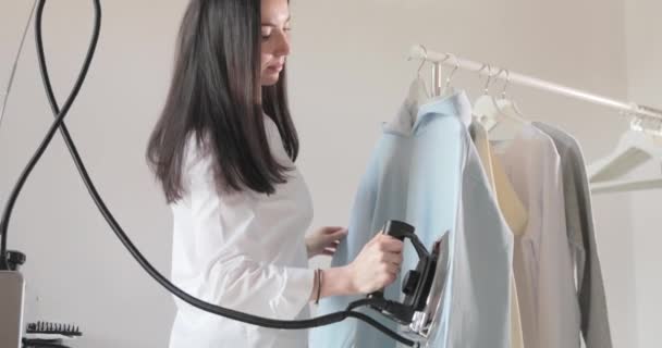 Αργή κίνηση βίντεο με το νεαρό όμορφο κορίτσι σιδερώνει ρούχα που κρέμεται στο κιγκλίδωμα, ατμούς σε κάθετη κατάσταση, ένα επαγγελματικό σίδερο με ατμό, ράψιμο παραγωγής, στούντιο, πρακτορείο — Αρχείο Βίντεο