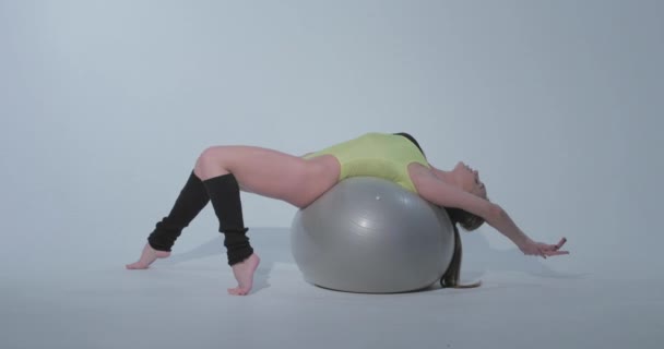 Mooi meisje in een geel sportpak en zwarte legging ligt op een bal, ze is bezig met gymnastiek op een grijze rubberen bal, badpak, mooie huid, figuur, blootsvoets, lang haar in de staart — Stockvideo