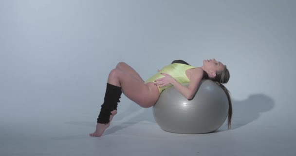 Beautiful girl in a yellow sports suit and black leggings lies on a ball, she is engaged in gymnastics on a gray rubber ball, swimsuit, beautiful skin, figure, barefoot, long hair in the tail — Stock Video