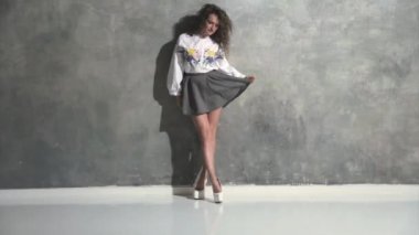 Kıvırcık saçlı, kıvırcık saçlı, kameranın önünde kıvrılıp flört eden, beyaz topuklu, kırmızı bir ayakkabı tabanı, kısa bir etek, gri bir arka plan, farklı duygular...