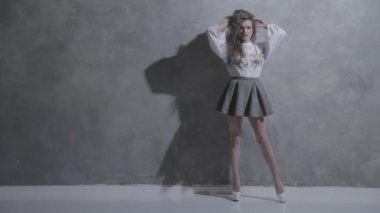 Kıvırcık saçlı, kıvırcık saçlı, kameranın önünde kıvrılıp flört eden, beyaz topuklu, kırmızı bir ayakkabı tabanı, kısa bir etek, gri bir arka plan, farklı duygular...