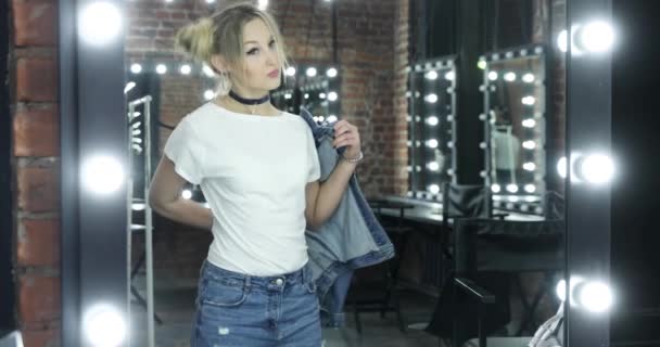 Het meisje de tiener in jeans jas, witte t-shirt poseert voor de spiegel in een make-up kamer, dansen, flirten, wriggles, een haarjurk in de vorm van bossen, een choker op de hals, armbanden op handen — Stockvideo