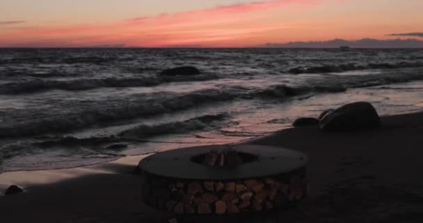 El paisaje del metal hermoso alrededor de una chimenea en la costa arenosa con un maremoto al atardecer, piedras, tranquilidad, el sol cae, leña, carbones rojos, nadie — Vídeos de Stock