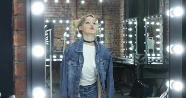Das Mädchen der Teenager in Jeansjacke, weißem T-Shirt posiert vor dem Spiegel in einem Schminkraum, tanzt, flirtet, zappelt, eine Frisur in Form von Trauben, einen Choker am Hals, Armbänder an den Händen — Stockvideo