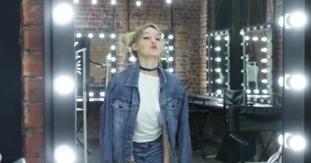 La fille l'adolescente en jean veste, t-shirt blanc pose devant le miroir dans une salle de maquillage, danses, flirts, tortillements, une coiffure en forme de grappes, un étranglement sur le cou, bracelets sur les mains — Video