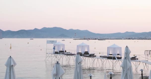 Franciaország, Cannes, 2017. május 27.: Cannes ünnepi töltése a Cannes-i Filmfesztiválon naplementekor, strandkávézók, esernyők, zsúfolt kávézók, rózsaszín ég — Stock videók