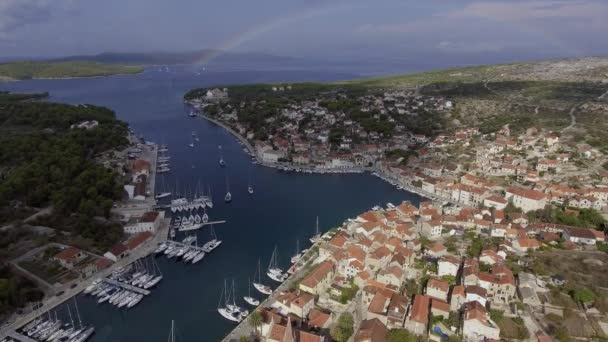 Вид с воздуха на порт в Мильне, Хорватия, точка обзора дрона, полная радуга на горизонте, острова, припаркованные яхты, рыбацкие лодки, домики с красными крышами — стоковое видео
