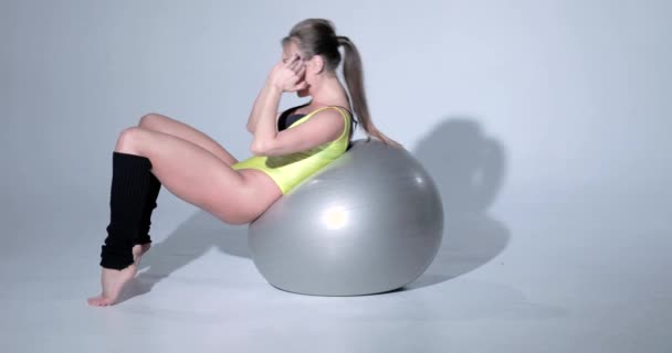 Beautiful girl in a yellow sports suit and black leggings lies on a ball, she is engaged in gymnastics on a gray rubber ball, swimsuit, beautiful skin, figure, barefoot, long hair in the tail — Stock Video