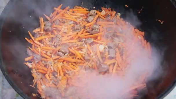 Przygotowanie krajowej uzbeckiej potrawy pilaf, pilaw, plov, marchew z mięsem na dużej patelni. Proces gotowania, otworzyć ogień. Gotowanie w płonącym kotle. Mieszaj powoli z odpieniaczem. Dodawanie drobno krojonej marchewki — Wideo stockowe