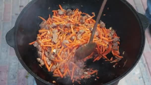 La preparación del plato nacional uzbek pilaf, pilaw, plov, la zanahoria con la carne en la cacerola grande. Proceso de cocción, fuego abierto. Cocinar en un caldero en llamas. Revuelva lentamente con un skimmer. Adición de zanahorias cortadas pequeñas — Vídeos de Stock