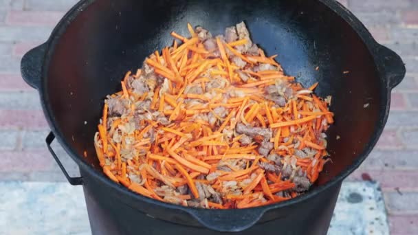 Preparação de prato uzbeque nacional pilaf, pilaw, plov, cenoura com a carne na grande panela. Processo de cozedura, fogo aberto. Cozinhar num caldeirão a arder. Mexa lentamente com uma escumadeira. Adição de cenouras de redução pequenas — Vídeo de Stock