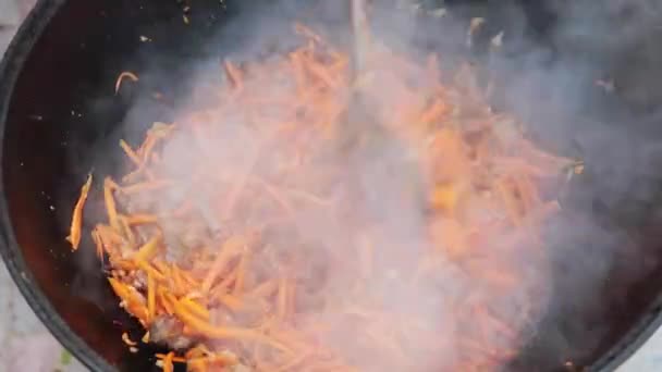 Persiapan hidangan nasional uzbek pilaf, pilaw, plov, wortel dengan daging di panci besar. Proses memasak, api terbuka. Memasak dalam kuali di atas api. Aduk perlahan-lahan dengan sebuah skimmer. Penambahan wortel potong kecil — Stok Video