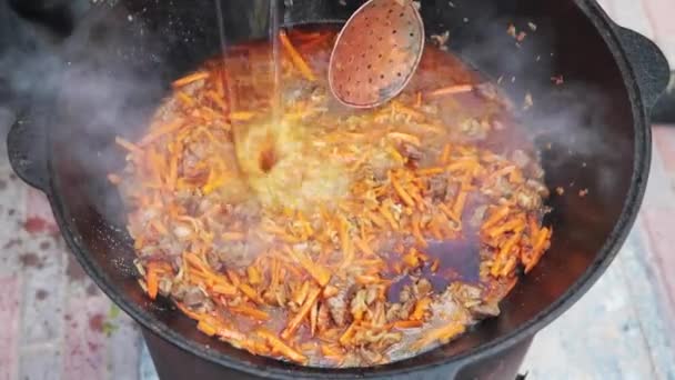 Preparation of national uzbek dish pilaf, pilaw, plov, carrot with meat in big pan. Add water, Cooking process, open fire. Cooking in a cauldron on fire. Stir slowly with a skimmer. Add spices — Stock Video