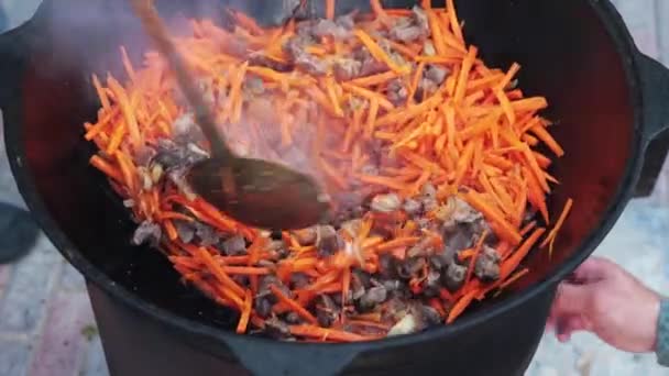 La preparación del plato nacional uzbek pilaf, pilaw, plov, la zanahoria con la carne en la cacerola grande. Proceso de cocción, fuego abierto. Cocinar en un caldero en llamas. Revuelva lentamente con un skimmer. Adición de zanahorias cortadas pequeñas — Vídeos de Stock