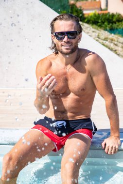 Güneş gözlüklü, uzun saçlı ve çıplak gövdeli yakışıklı adam havuz kenarında oturur, su sıçrar, eğlenir, bronz tenli spor yapar, kırmızı mavi yüzme şortu, güneşli bir gün