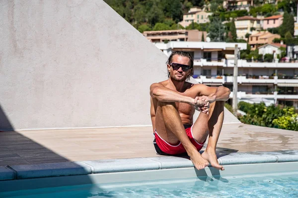 The handsome brutal man in sunglasses with a long hair and naked torso sits near the pool, is looking a camera, a sports suntanned body, sunglasses with a blue frame, he is red blue swimming shorts