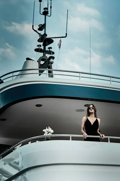 Гламурная дива в вечернем платье черного цвета и солнечных очках стоит на верхней палубе огромной яхты в предвкушении — стоковое фото