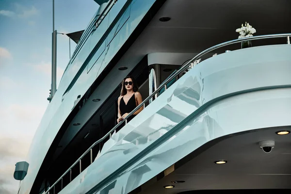 Okouzlující diva ve večerních šatech černé barvy a sluneční brýle stojí na horní palubě obrovské jachty v očekávání — Stock fotografie