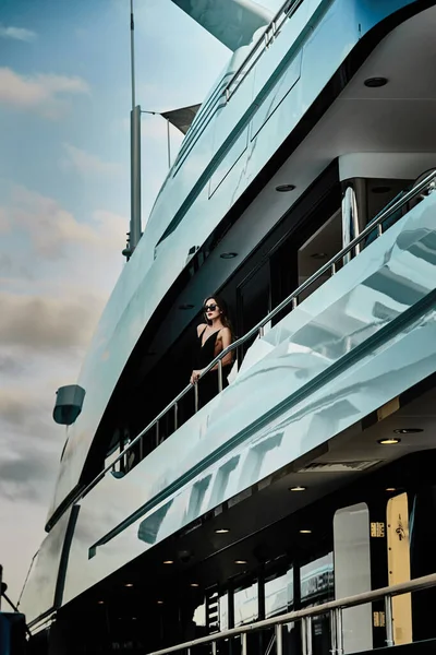 Гламурная дива в вечернем платье черного цвета и солнечных очках стоит на верхней палубе огромной яхты в предвкушении — стоковое фото