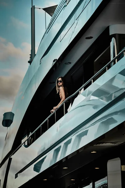 A glamorous diva in an evening dress of black color and sunglasses stands on the top deck of a huge yacht in anticipation — Stock Photo, Image
