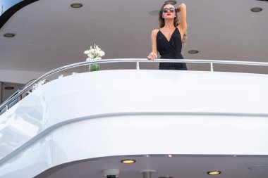 Siyah bir gece elbisesi ve güneş gözlüğü takmış zarif kız büyük bir yatın üst güvertesinde bekliyor. Kırmızı dudaklı, muhteşem bir kadın.