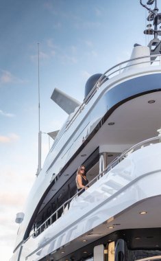 Siyah bir gece elbisesi ve güneş gözlüğü takmış zarif kız büyük bir yatın üst güvertesinde bekliyor. Kırmızı dudaklı, muhteşem bir kadın.