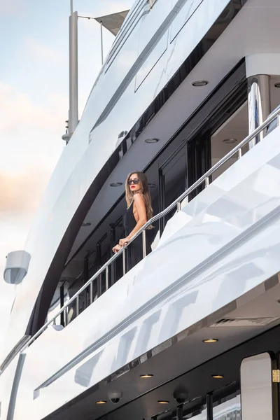 Az elegáns lány öltözött estélyi ruha fekete színű és napszemüveg áll a felső fedélzeten egy hatalmas jacht várakozás, piros ajkak, gyönyörű hölgy, ő nem fel haj — Stock Fotó