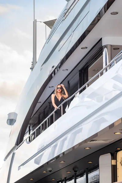 La chica elegante vestida con un vestido de noche de color negro y gafas de sol se encuentra en la cubierta superior de un enorme yate en anticipación, labios rojos, hermosa dama, ella hace el pelo — Foto de Stock