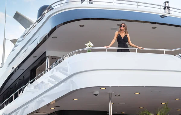 The elegant girl dressed in an evening dress of black color and sunglasses stands on the top deck of a huge yacht in anticipation, red lips, gorgeous lady, she does up hair — Stock Photo, Image