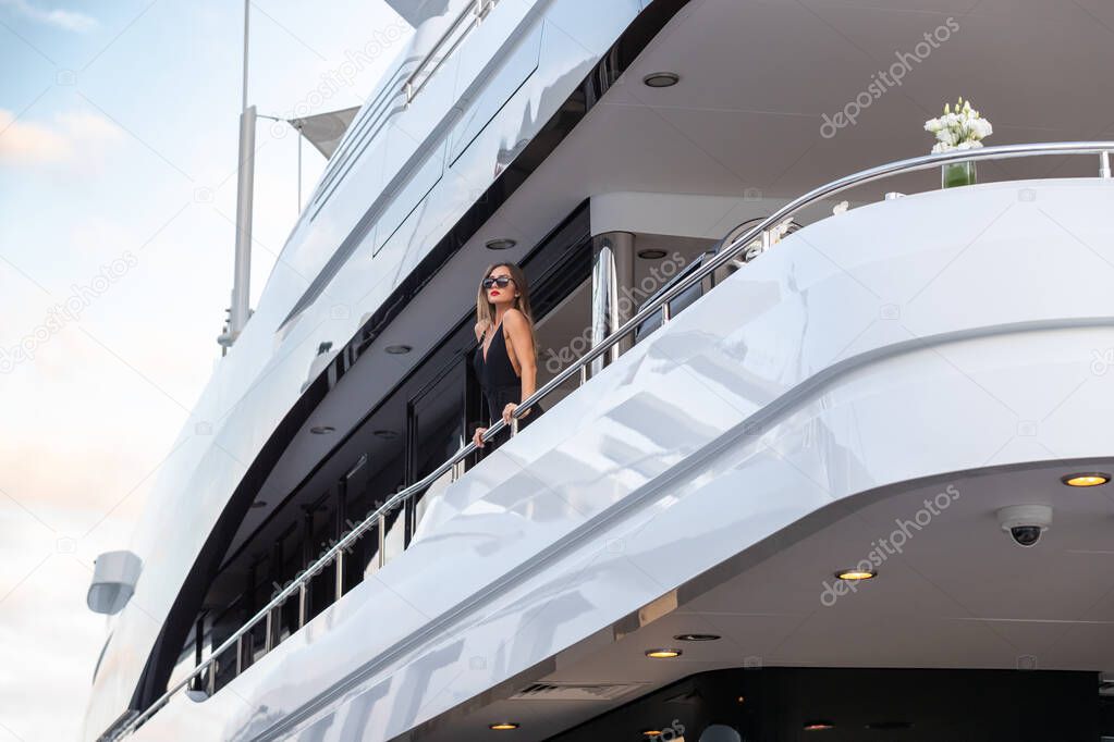 The elegant girl dressed in an evening dress of black color and sunglasses stands on the top deck of a huge yacht in anticipation, red lips, gorgeous lady, she does up hair