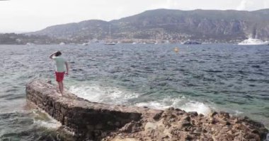 Güney Fransa, Fransız Rivierası, kırmızı şortlu ve yeşil tişörtlü yakışıklı genç adam küçük bir iskelede, deniz sıçramaları, arka planda liman ve dağlar, dalgalar, yatlar., 