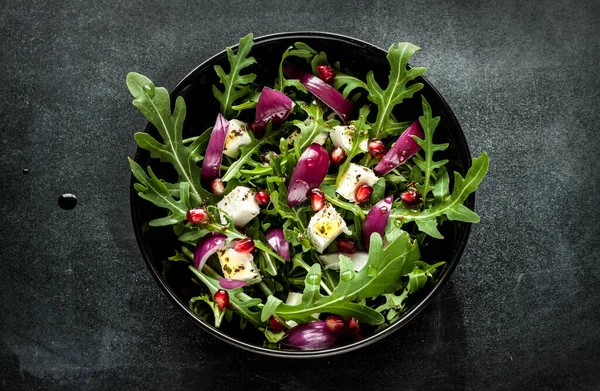 Fresh spring salad with rucola, feta cheese, red onion and pomegranate seeds in black bowl on chalkboard background. Captured from above (top view).