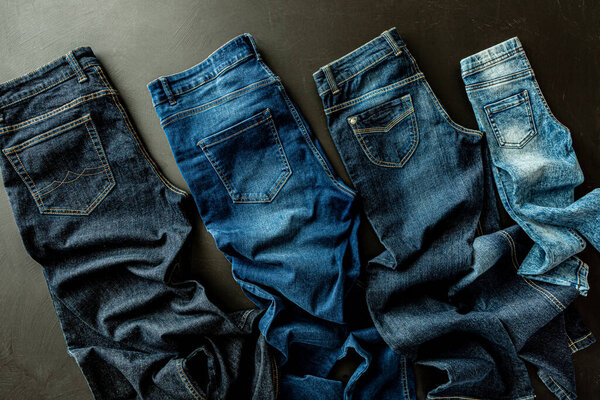 Clothing - family jeans on black chalkboard background. Denim trousers captured from above (top view, flat lay). Various sizes, shapes and blue shades (tones).