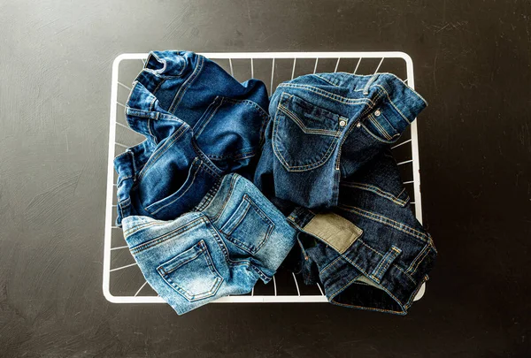 Family jeans in laundry (washing) basket on black chalkboard background. Denim trousers captured from above (top view). Various sizes, shapes and blue shades (tones).