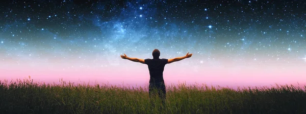 Человек с распростертыми руками стоит на травяном поле против ночного звездного неба. Элементы этого изображения предоставлены НАСА . — стоковое фото