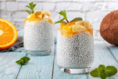 Mavi ahşap bir masada, çiğ chia tohumlu iki cam bardakta muzlu chia pudingi.