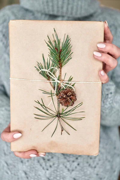 Girl holds a book for a gift wrapped in craft paper decorated with a fir branch with a cone