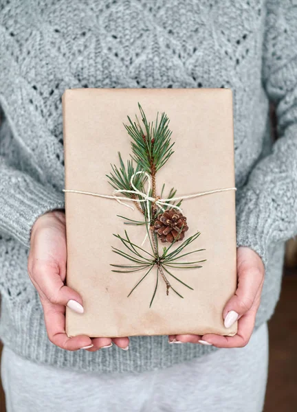 Girl holds a book for a gift wrapped in craft paper decorated with a fir branch with a cone