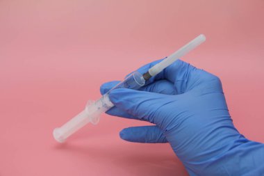 Mavi eldivenli bir elin elinde şırınga var. Pembe arka plan. Hastalık tedavisi konsepti. Vücudu virüsten koruyorum. Aşılama. Doktor aşı için bir şırınga tutuyor. İlaç..