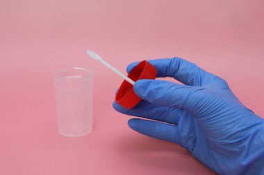 Mavi lateks eldivenli bir el dışkı toplamak için bir kap tutar. Doktorun elinde analiz için plastik kap var. Analiz kavanozu boş. Pembe arkaplan.