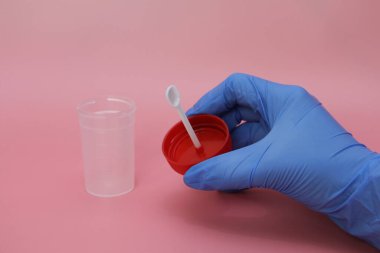 Mavi lateks eldivenli bir el dışkı toplamak için bir kap tutar. Doktorun elinde analiz için plastik kap var. Analiz kavanozu boş. Pembe arkaplan.