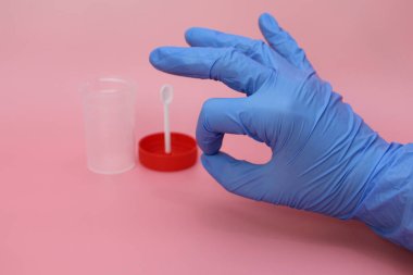 Mavi lateks eldivenli bir el dışkı toplamak için bir kap tutar. Doktorun elinde analiz için plastik kap var. Analiz kavanozu boş. Pembe arkaplan.