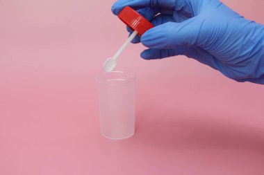 Mavi lateks eldivenli bir el dışkı toplamak için bir kap tutar. Doktorun elinde analiz için plastik kap var. Analiz kavanozu boş. Pembe arkaplan.