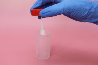 Mavi lateks eldivenli bir el dışkı toplamak için bir kap tutar. Doktorun elinde analiz için plastik kap var. Analiz kavanozu boş. Pembe arkaplan.